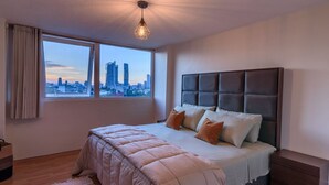Un dormitorio con vista a la ciudad, una cama grande con muchas almohadas, cabecera y lámpara colgante.