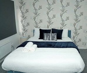 A neatly made bed with white bedding and a dark blue accent, two rolled towels, and a blue headboard. - Family & Contractor Haven | Sleeps 6 | Parking & Garden (West Midlands)