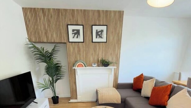 Una sala moderna con un televisor de pantalla plana, un sofá gris con cojines naranja, una mesa redonda y una pared de acento de madera.