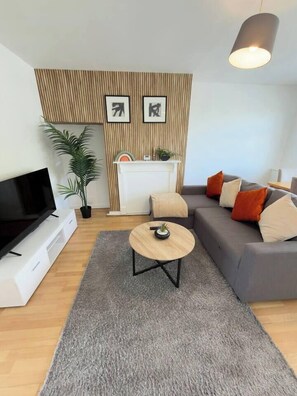 Ein modernes Wohnzimmer mit einem Flachbildfernseher, einem grauen Sofa mit orangefarbenen Kissen, einem runden Couchtisch und einer Wandvertäfelung aus Holz.