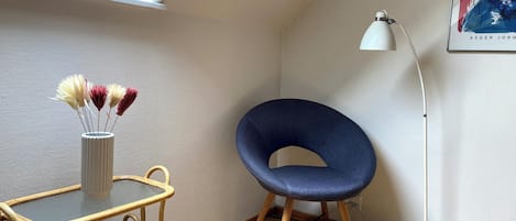Una esquina de una habitación con una silla azul, una lámpara de pie blanca, un jarrón blanco con flores secas y una mesa de mimbre con libros.