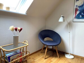 A corner of a room with a blue chair, a white floor lamp, a white vase with dried flowers, and a wicker table with books.