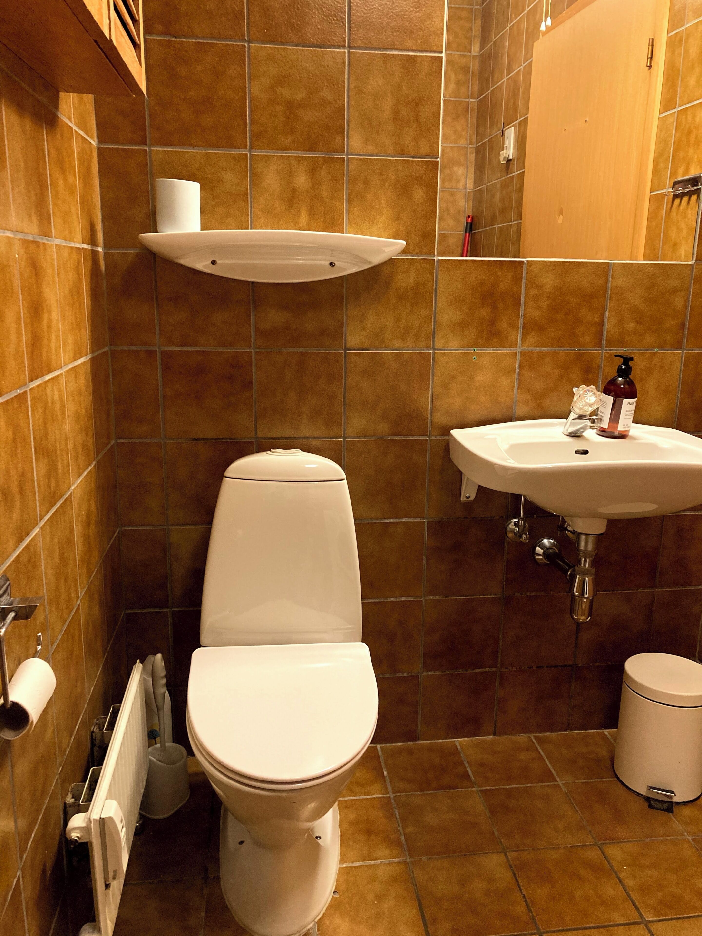 Et badeværelse med toilet, håndvask og et hyldebord over toilettet.
