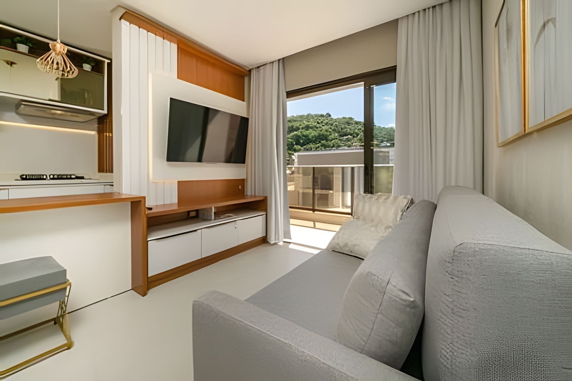 A modern living room with a grey sofa, a flat-screen TV mounted on a wooden cabinet, a dining table with a gold frame, and a balcony with a view of greenery.