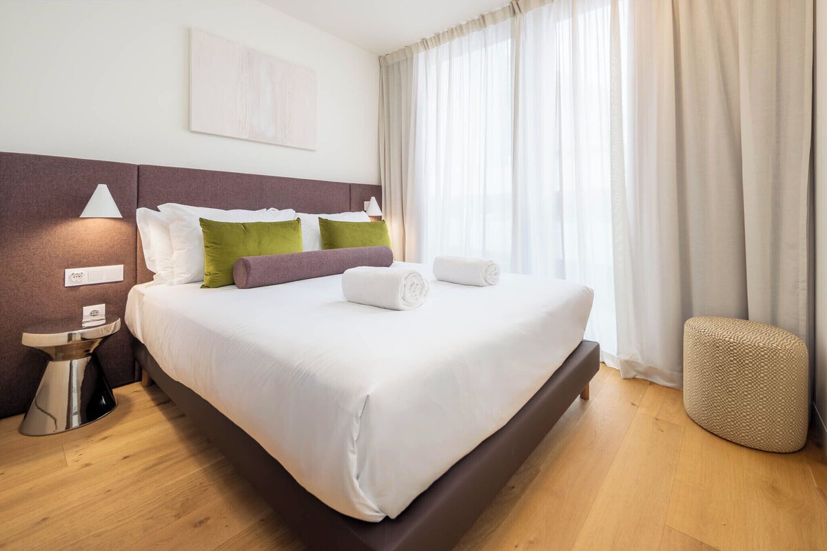 A hotel room with a large bed, a bedside lamp, a small table, and a wall-mounted light fixture.