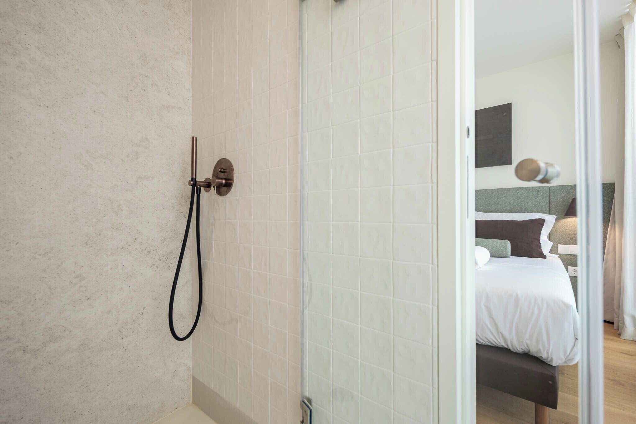 A modern bathroom with a shower, a glass door leading to a bedroom with a bed and a nightstand.