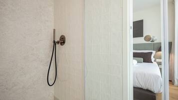 A modern bathroom with a shower, a glass door leading to a bedroom with a bed and a nightstand.