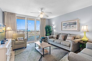 A living room with a sofa, armchair, coffee table, and a view of the ocean through large windows. - Pelican Beach Terrace 1503 (Destin)