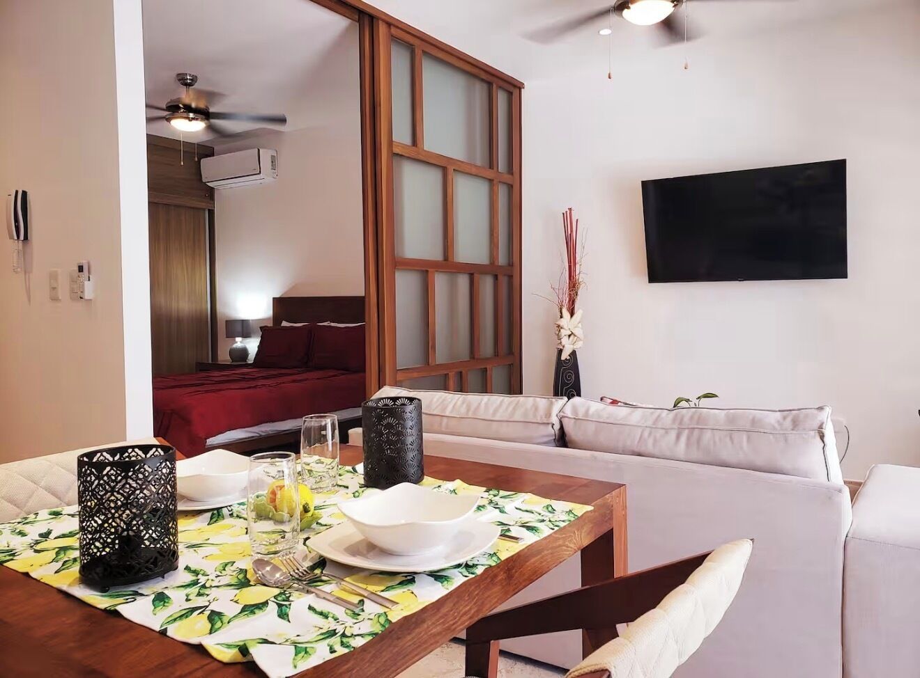 A dining area with a table set for two, a sofa, a TV mounted on the wall, and a ceiling fan.