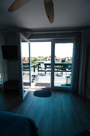 Habitación de hotel con cama, ventilador de techo, televisor y puerta corrediza de vidrio que da a un espacio exterior con mesa y sillas.