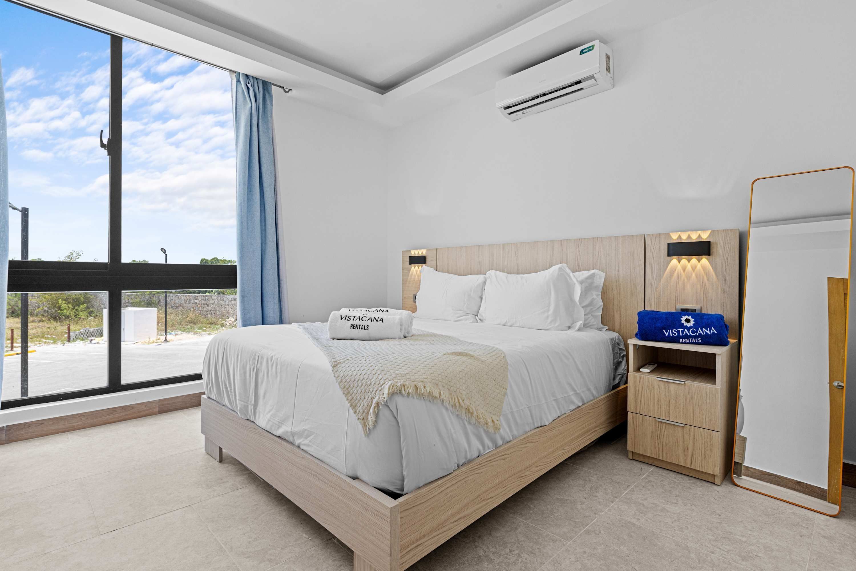 A modern bedroom with a large bed, a nightstand, a mirror, and an air conditioning unit.