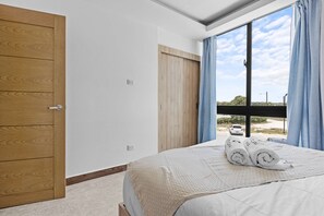 A bedroom with a bed, a wooden wardrobe, a window with curtains, and a view of a parking area. - NOK Caribbean 1BR Retreat By Vistacana (Punta Cana)