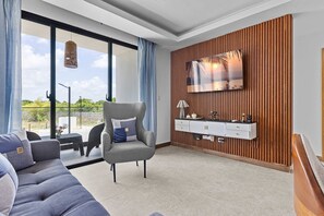 Ein modernes Wohnzimmer mit grauem Sofa, einem einzelnen Sessel, einem an einer Holzvertäfelung befestigten Fernseher und einem Balkon mit Blick auf Grünflächen.