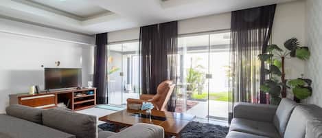 A modern living room with a flat-screen TV, a wooden entertainment unit, a grey sofa, a coffee table, and a large window with curtains.