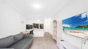 Um espaço de estar moderno com um sofá cinzento, uma área de cozinha e uma grande obra de arte com tema de praia na parede.