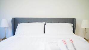 Cama bem arrumada com lençóis brancos, cabeceira cinza e duas luminárias laterais.