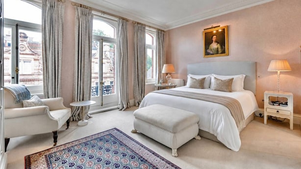 A hotel room with a large bed, a sitting area with a chair and a small table, a painting on the wall, and a view through the window. - Elegant Mayfair Home Central AC Mount Street (London)