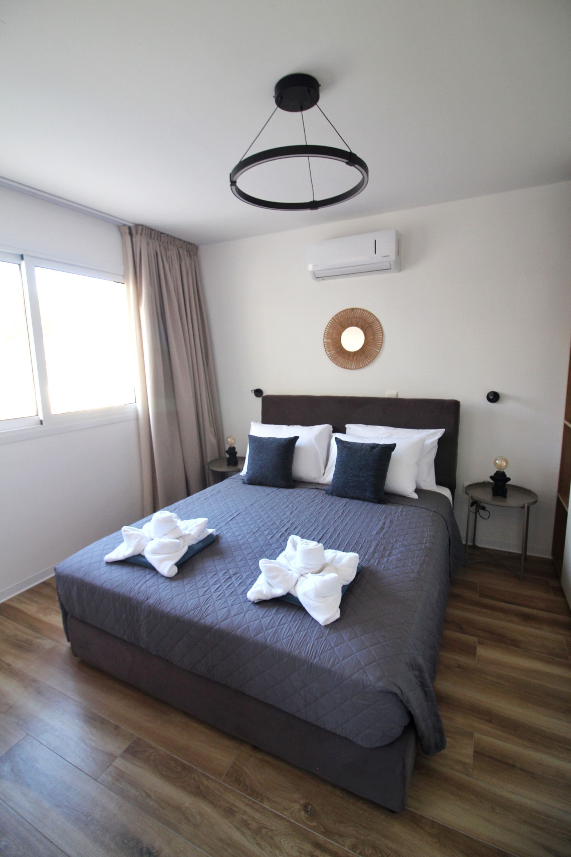 A hotel room with a large bed, a round wall mirror, an air conditioning unit, and a window with curtains.
