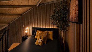 Una habitación con techo inclinado, paneles de madera en las paredes, una cama con ropa de cama negra y varias almohadas, una mesita de noche con una lámpara y una planta grande en maceta.