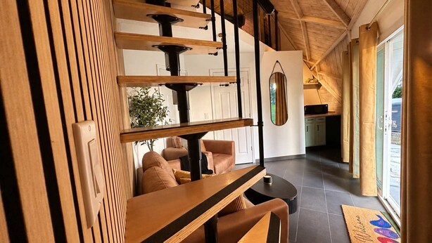 Un interior moderno con una escalera de madera, una zona de estar con un sofá y sillones, y una cocina visible a través de una puerta abierta.