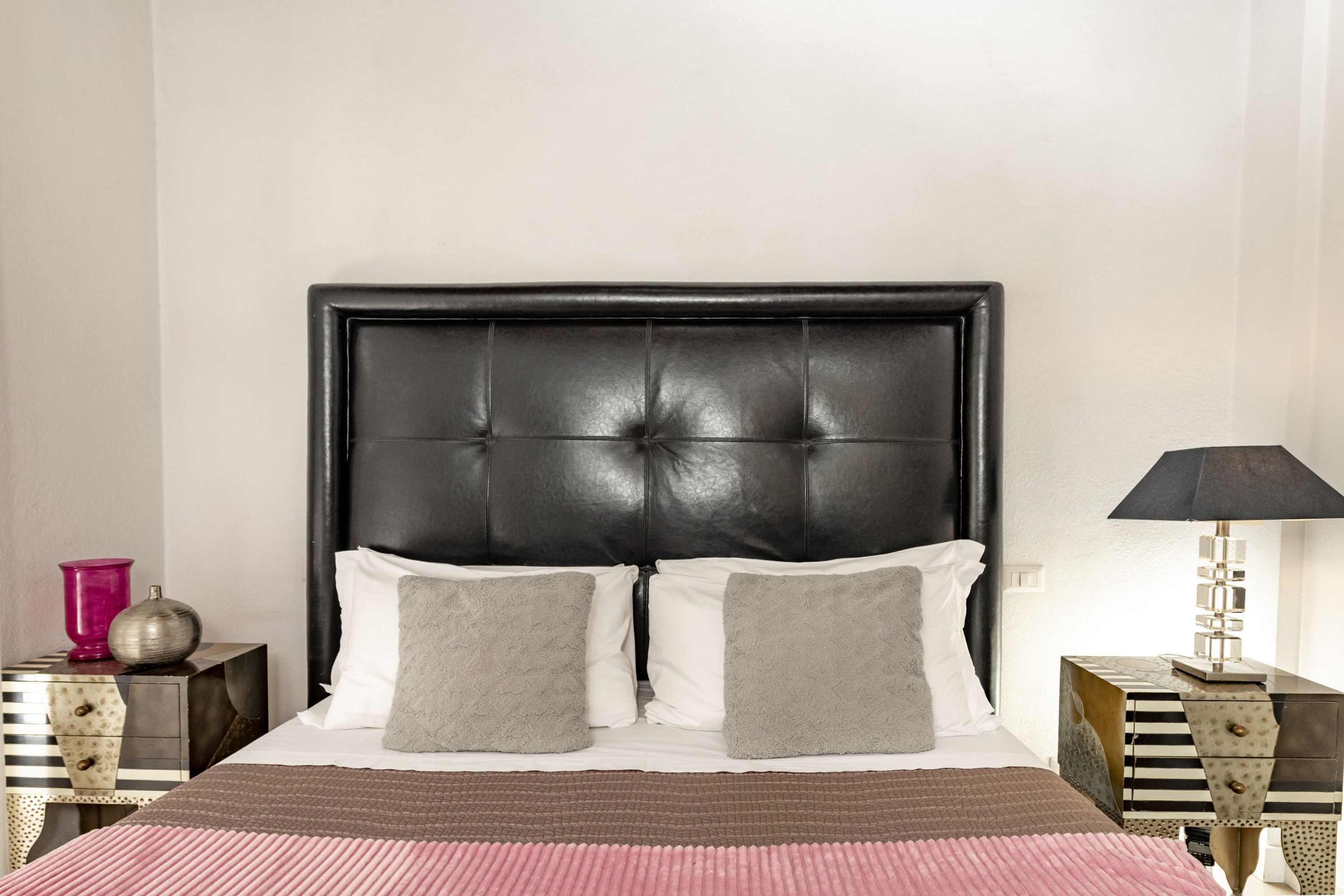 Una cama con cabecera capitonada, dos almohadas, una colcha estampada y una mesita de noche con una lámpara.