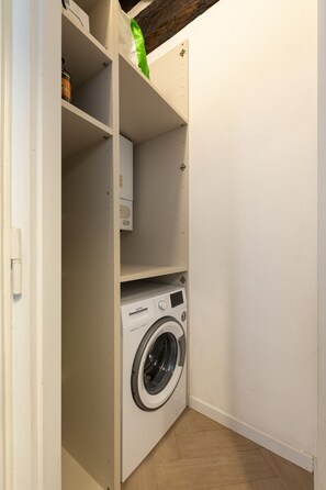 A compact laundry room with a front-loading washing machine, a shelf with a green item, and a white door. - San Francesco Ripa 4 - Lorenza 3BR Group Family Home (Rome)