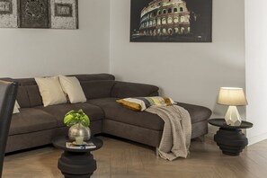 A living room with a sectional sofa, a small side table with a plant, a lamp, and a framed picture of a building. - San Francesco Ripa 4 - Lorenza 3BR Group Family Home (Rome)