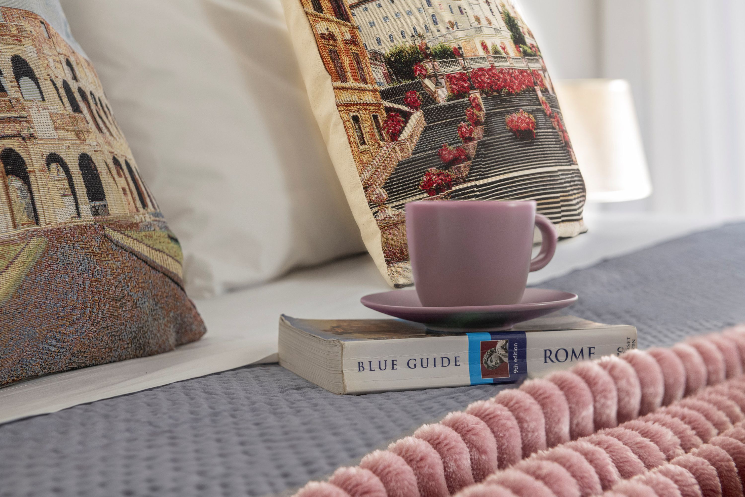 A bed with a grey textured blanket, a pink mug on a book titled 'BLUE GUIDE ROME,' and patterned pillows.