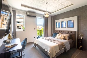 Un dormitorio moderno con una cama, mesitas de noche, un escritorio y una silla. Hay un balcón con vistas a edificios y una montaña.