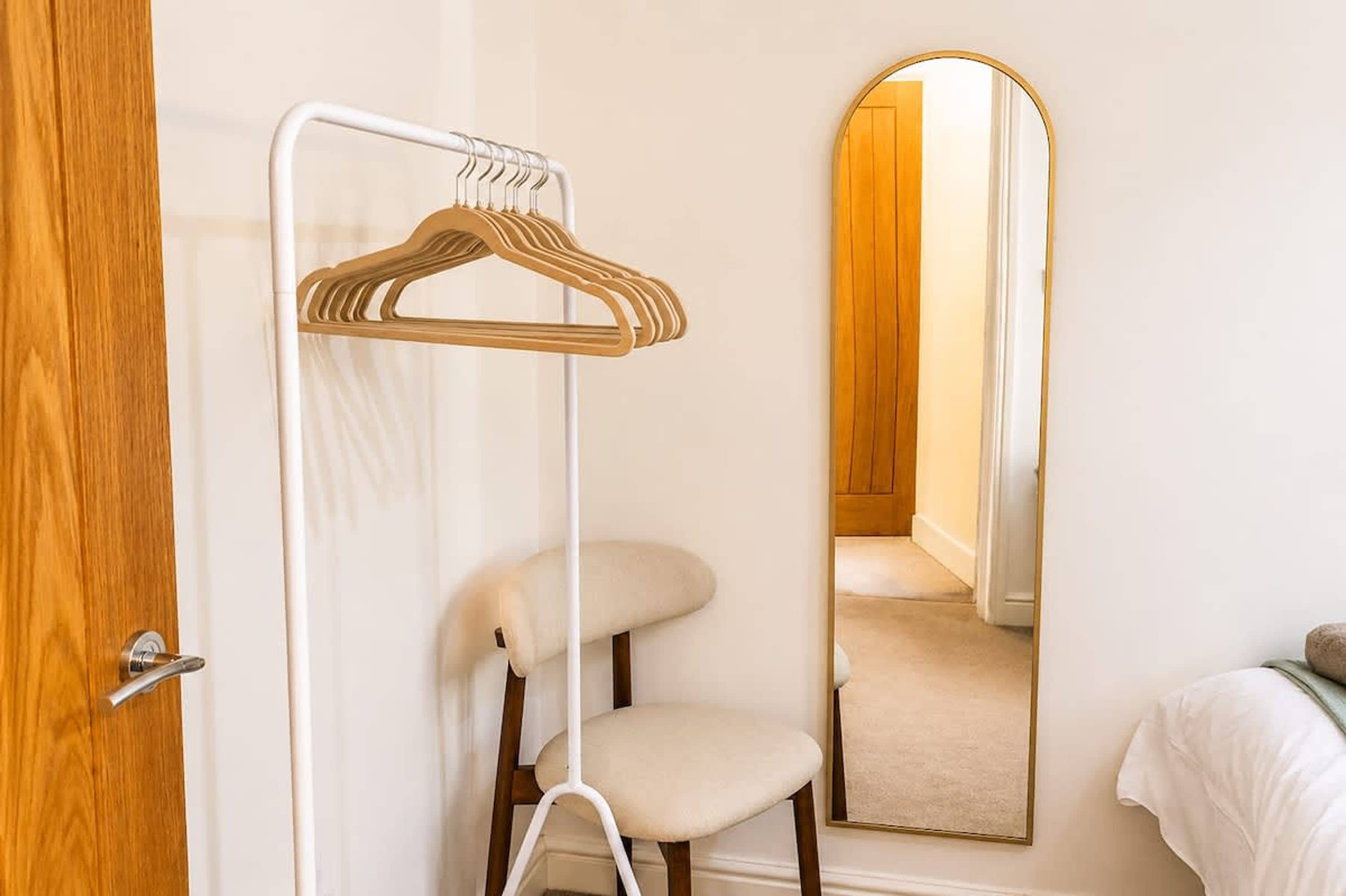 A room with a wooden door, a white wall-mounted hanger, a white chair, and a mirror reflecting a door and a hallway.