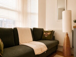 A green sofa with a white throw blanket and a bee pillow, a tall floor lamp, and a window with blinds. - Perfect Booking Bright 2 Bed Maisonette in Bath (Bath)