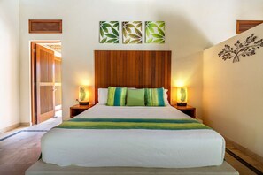 Ein Hotelzimmer mit einem hölzernen Kopfteil, einem Bett mit weiß-grünen Streifen, zwei Nachttischlampen und einer Wand mit Wandbildern im Blattmuster.