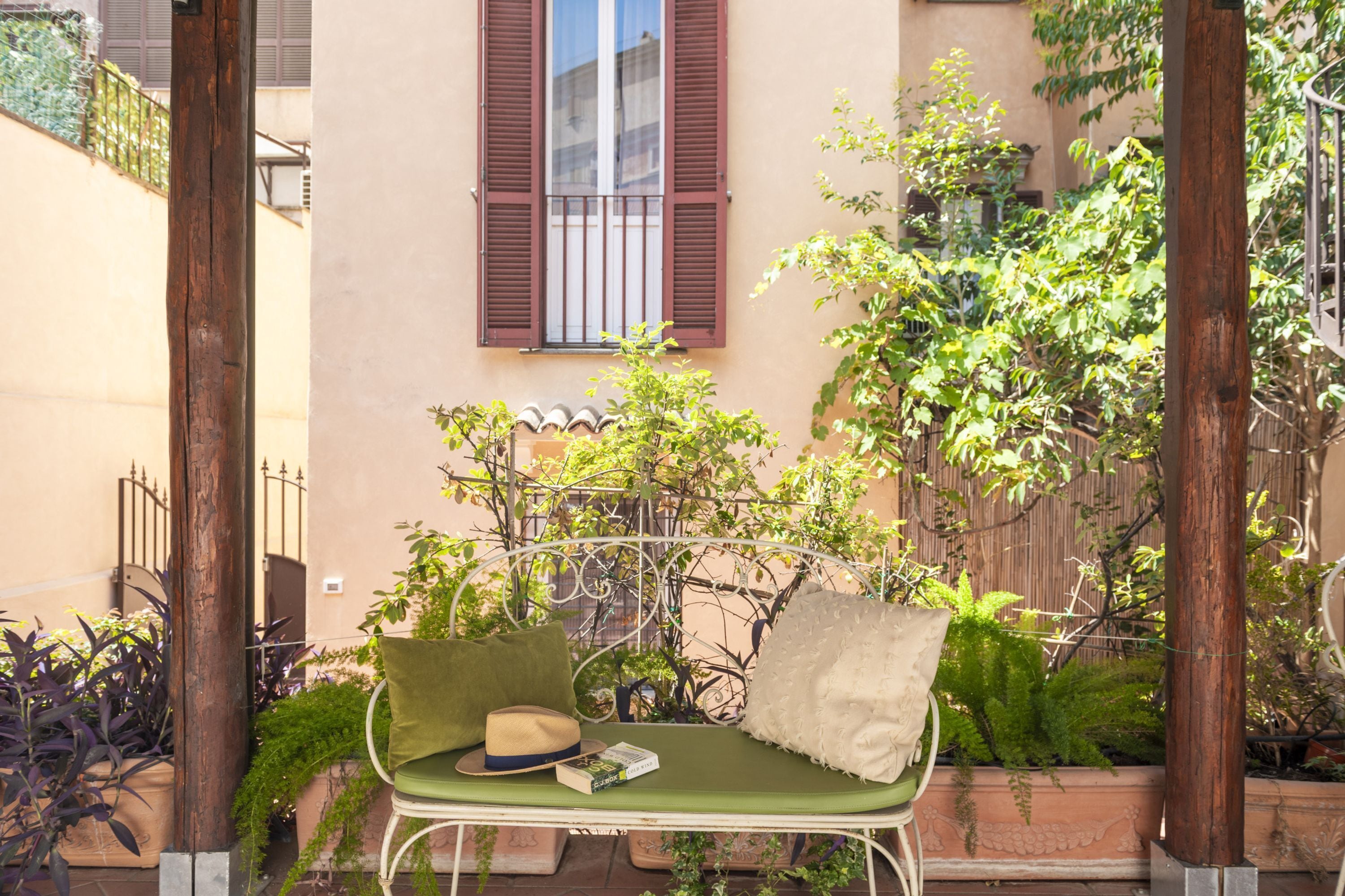 A green metal bench with cushions and a hat, positioned in a garden area with potted plants and a building in the background.