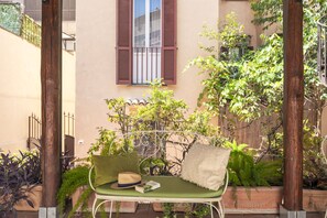 Un banco de metal verde con cojines y un sombrero, ubicado en un jardín con plantas en macetas y un edificio al fondo.
