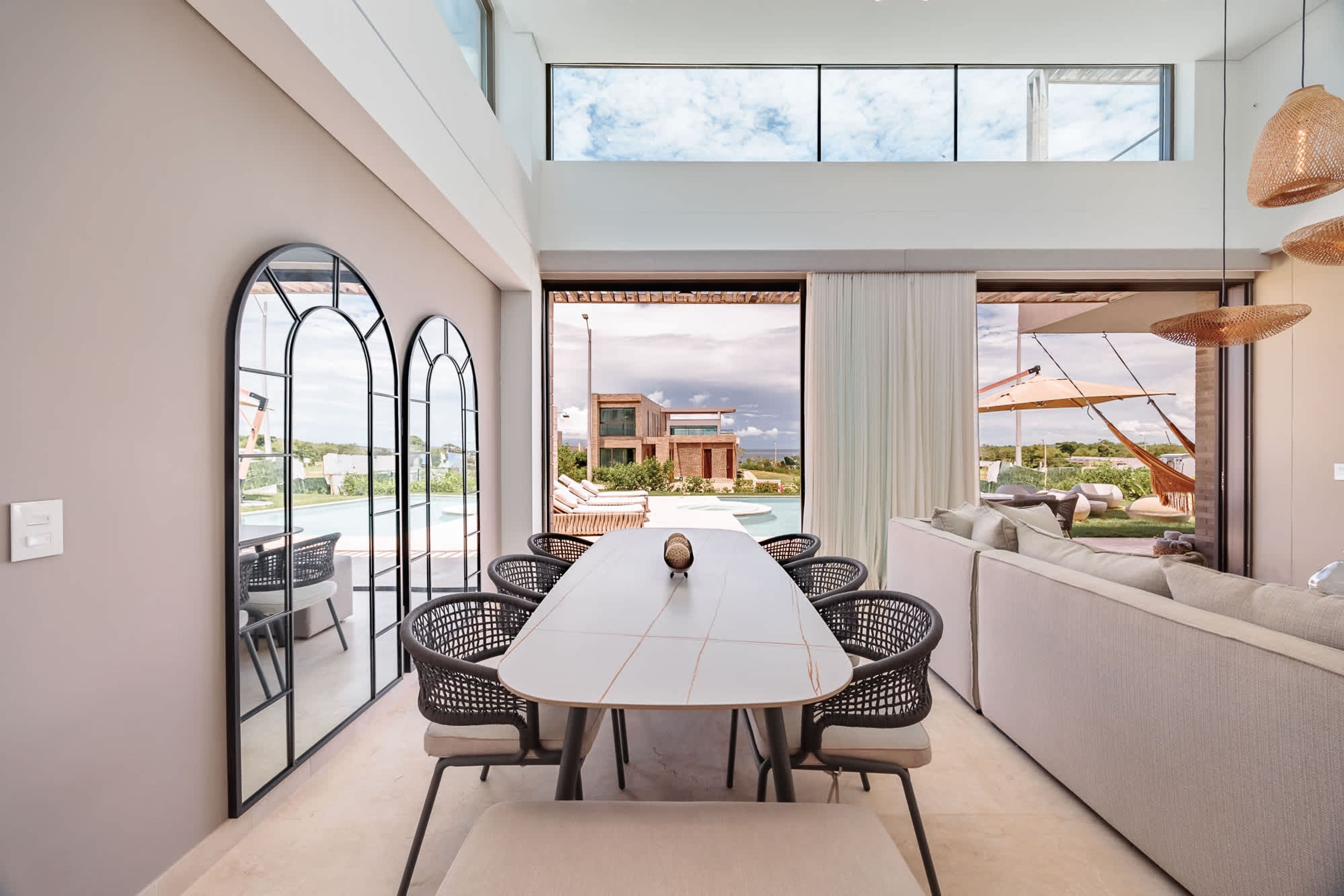 A modern dining area with a long table, black wicker chairs, and a view of a pool and building.