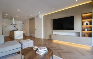 A modern living room with a sofa, ottoman, and coffee table, a kitchen area, and a large flat-screen TV mounted on a wall. - w Enjoyable 2BR Stay w Balcony in Miraflores (Lima)