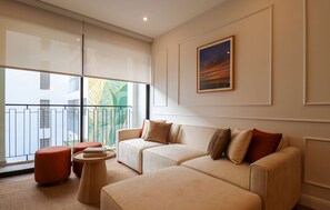 Ein modernes Wohnzimmer mit einem beigefarbenen Sofa, passendem Hocker und einem kleinen runden Tisch mit Büchern. Ein großes Fenster bietet Blick auf einen Balkon mit Geländer und ein Gebäude im Hintergrund.