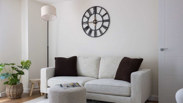 Ein Wohnzimmer mit einem hellgrauen Sofa, zwei dunkelgrauen Kissen, einer runden Wanduhr, einer Stehlampe, einem kleinen Beistelltisch, einer Topfpflanze und einer Zeitschrift auf dem Hocker.