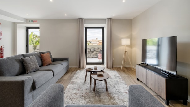 Uma sala de estar moderna com um sofá cinzento, uma mesa de centro, uma TV sobre um suporte de madeira e vista para o exterior através de uma porta de vidro.