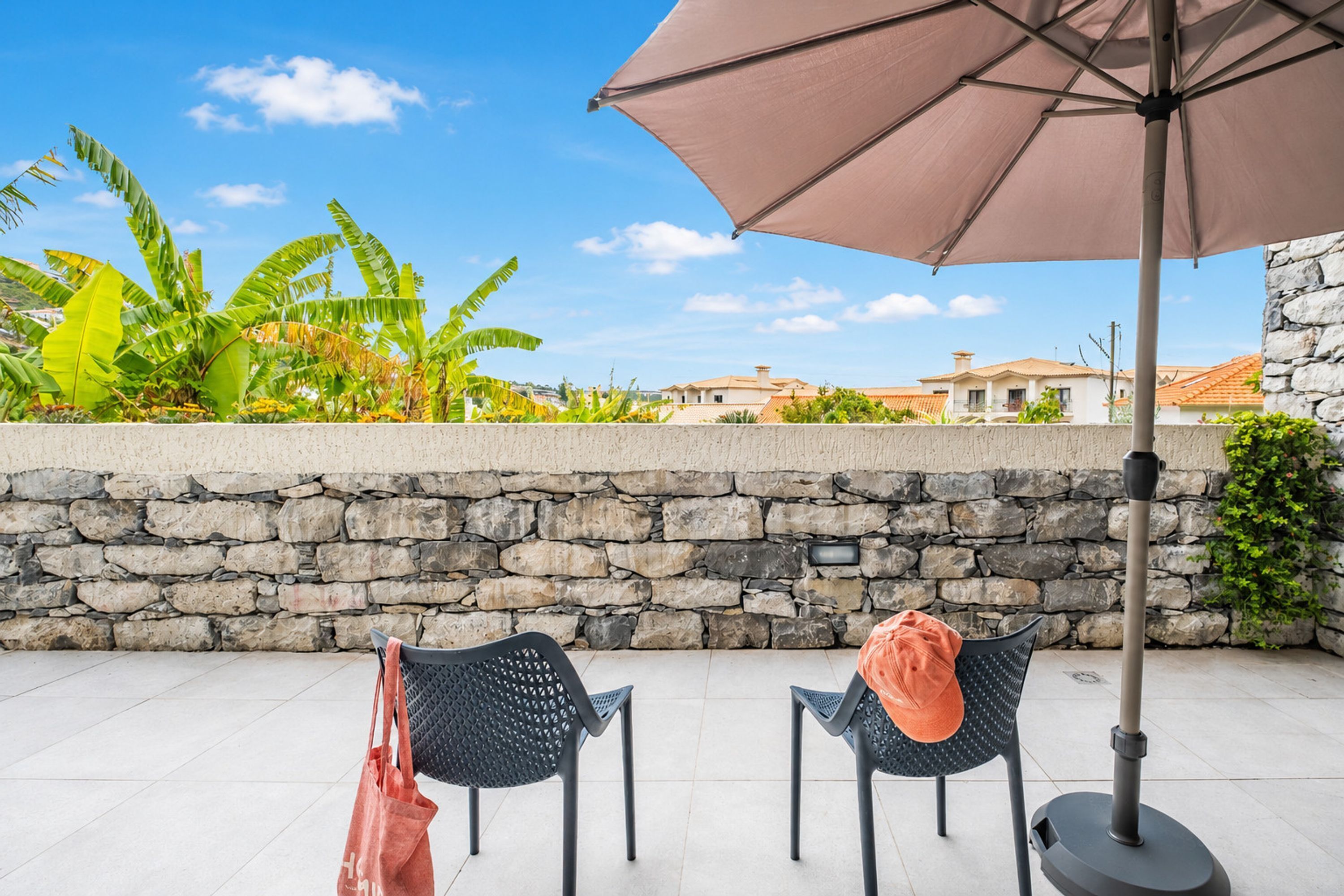 An outdoor seating area with two chairs and an umbrella, a stone wall, and a view of houses and greenery.