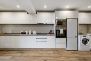 A modern kitchen with white cabinets, a built-in oven, microwave, and a washing machine. - Oceanea Apartment by Homie (Santa Cruz)