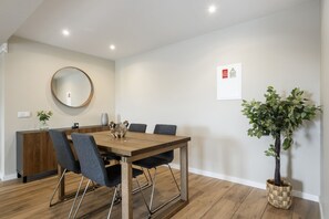 A modern dining area with a wooden table, grey upholstered chairs, a round mirror, and a potted plant. - Oceanea Apartment by Homie (Santa Cruz)
