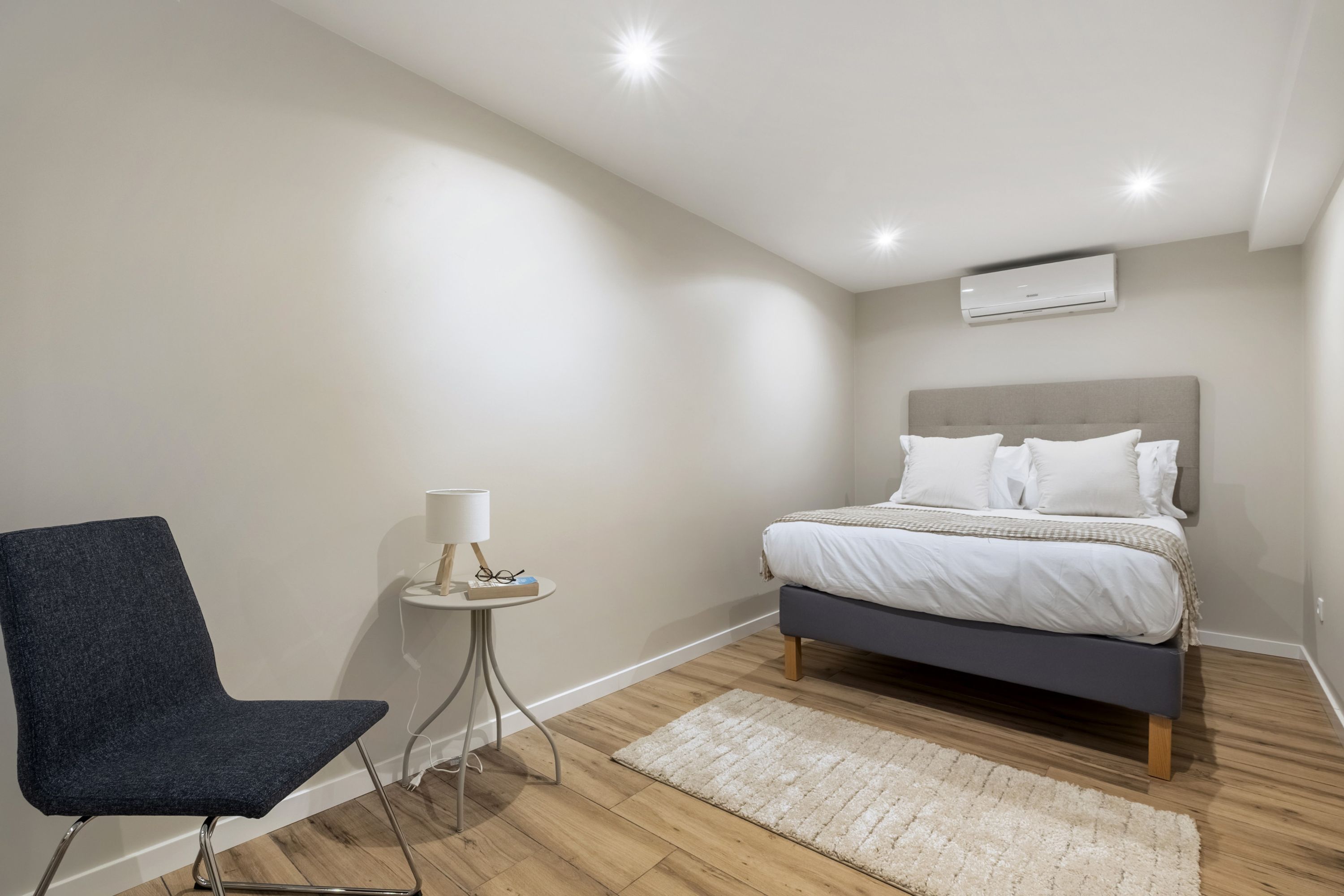 A bedroom with a bed, a chair, a small table with a lamp, and an air conditioning unit on the wall.