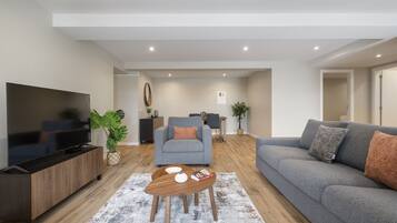 Una sala de estar moderna con una televisión de pantalla plana, sofás grises, una mesa de centro y un comedor al fondo.