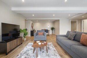 A modern living room with a flat-screen TV, grey sofas, a coffee table, and a dining area in the background.