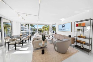 A modern living room with a glass dining table, sofas, a coffee table, and a bookshelf. - Chic Bay Harbor 3BR Rooftop Pool Free Parking (Bay Harbor Islands)