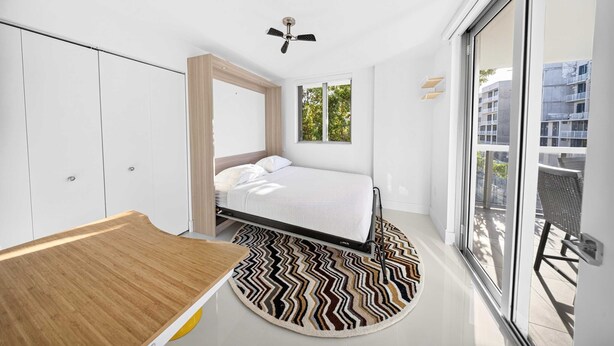 A modern bedroom with a bed, a wooden desk, a ceiling fan, and a large window offering a view of buildings. - Chic Bay Harbor 3BR Rooftop Pool Free Parking (Bay Harbor Islands)