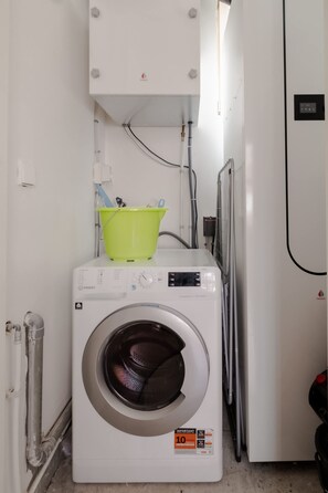 A compact laundry room with a front-loading washing machine, a green bucket, and a water heater. - In het Hart van Schiedam Rotterdam om de Hoek (Schiedam)