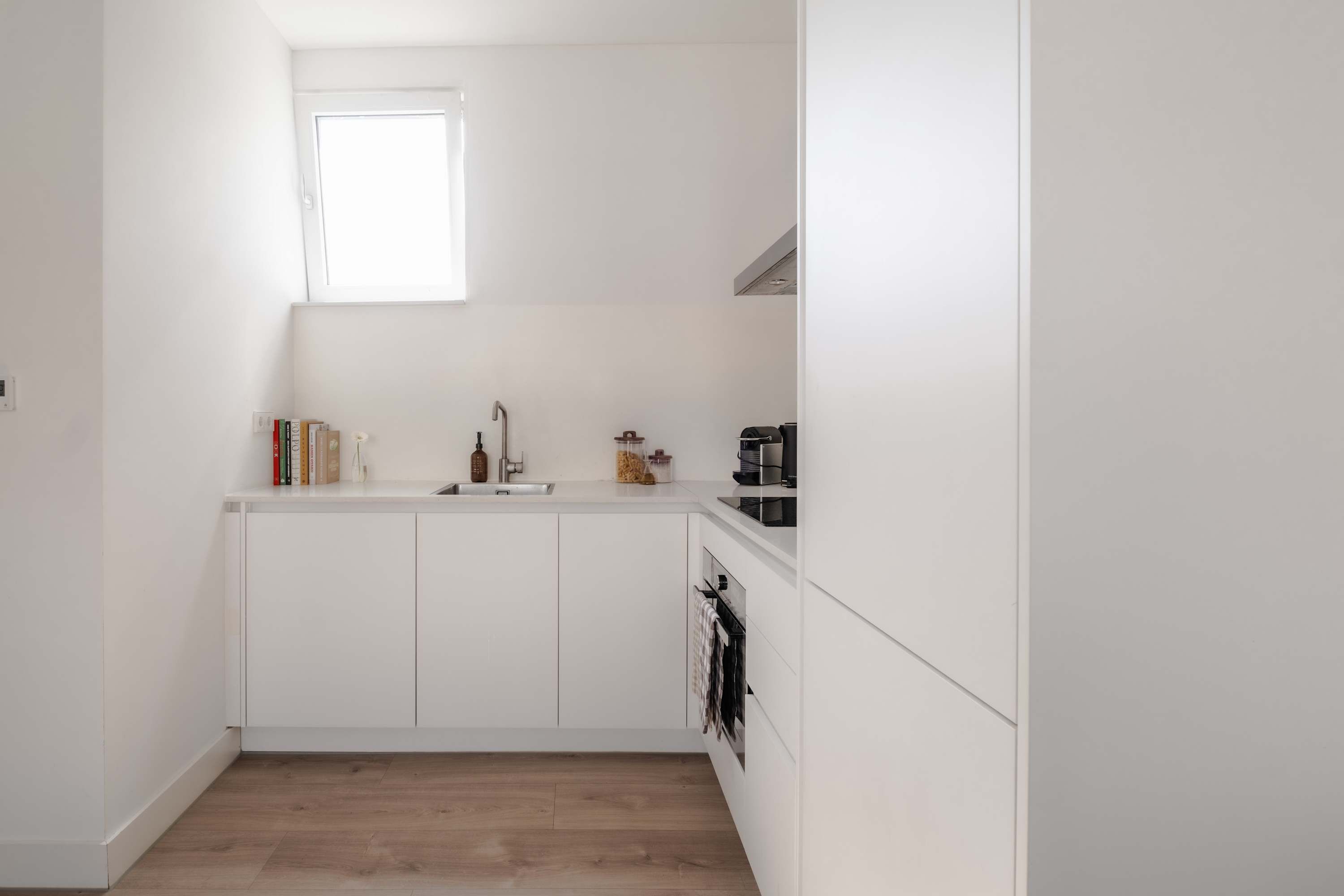 A small, white kitchen with a window above the sink, a coffee machine, and a shelf with books and a vase.