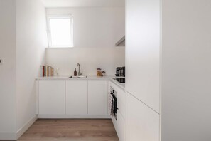 A small, white kitchen with a window above the sink, a coffee machine, and a shelf with books and a vase. - In het Hart van Schiedam Rotterdam om de Hoek (Schiedam)